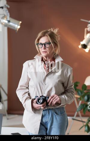 Portrait von reifen Fotografen in Brillen halten Fotokamera und Blick auf die Kamera, während Sie im Studio stehen Stockfoto
