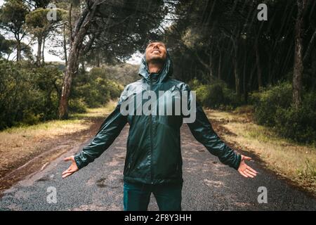Junger Mann mit nassem Lächeln in einer durchnässten Jacke mit Kapuze, der den Regen genießt. Mann im Regenmantel stand bei starkem Regen. Schlechter Herbst, egal. Stockfoto