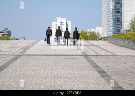 Vier japanische Salarymen in schwarzen Business-Anzügen gehen von der Kamera auf dem Gehweg in der Tokio-Bucht, Japan, weg Stockfoto