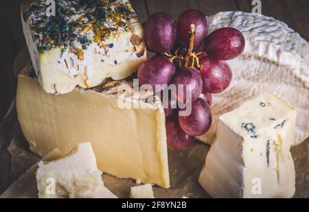 Auswahl an exklusivem Käse, dekoriert mit roten Trauben, auf einem rustikalen Holztisch, Nahaufnahme Stockfoto