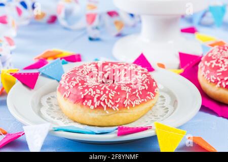 Pinkfarbener glasierter Donut auf einem weißen Teller auf hellblauem Hintergrund, verziert mit einem Streamer und einer Partygirlande Stockfoto