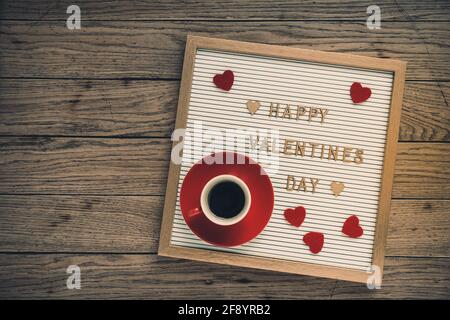 Weiße Tafel mit Goldschriftzug für den Valentinstag, rote Herzen und eine rote Tasse Espresso auf rustikalem Holzhintergrund, Draufsicht Stockfoto