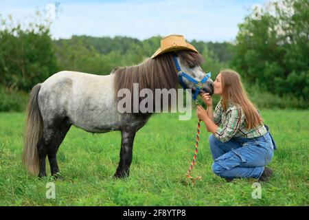 Die junge kaukasische Frau sitzt auf dem Gras und küsst ihr Pferd in der Schnauze, Seitenansicht. Stockfoto