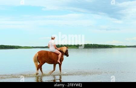 Eine kaukasische Frau reitet auf ihrem roten Pferd entlang des Wassers in einer flachen Lagune. Stockfoto