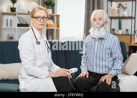 Porträt eines älteren grauhaarigen bärtigen Patienten mit Vernebler-Inhalator-Maske, sitzend mit seinem lächelnden professionellen Arzt und schauend in die Kamera. Arzt, der den Patienten zu Hause zur Inhalation besucht. Stockfoto