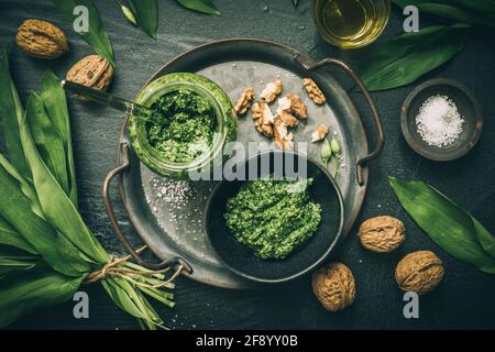 Hausgemachtes Bärlauch-Pesto auf einem Metalltablett, Bärlauch-Blätter, Salz, Öl und Walnüsse auf dunkelgrünem Hintergrund, Draufsicht Stockfoto