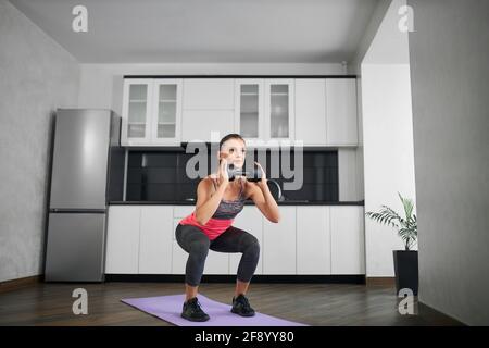 Seitenansicht einer jungen kaukasischen Frau, die in der Küche hockt. Starkes Mädchen trägt Sportkleidung Trainingsbeine auf Matte zu Hause am Morgen trägt Hantel in den Händen. Konzept der Home-Workout, Gewichtsverlust. Stockfoto