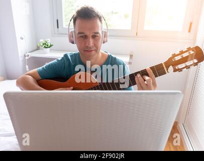 Kaukasischer Mann überprüft seinen Laptop, während er Gitarre spielen lernt Online Stockfoto