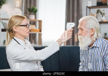 Selbstbewusste blonde Ärztin im Alter von 40 Jahren, die die Körpertemperatur eines älteren bärtigen Patienten mit berührungslosen Infrarot-Stirnthermometern während des Hausbesuchs überprüft. Stockfoto