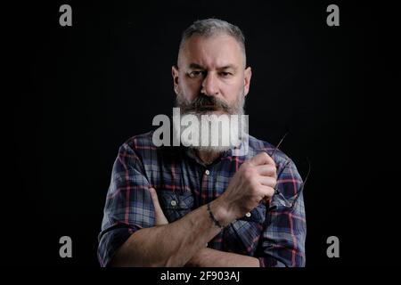 Studioportrait eines reifen bärtigen Mannes in legerer Kleidung mit Brille in der Hand, der direkt auf die Kamera blickt, zwangloser Lifestyle, schwarzer Hintergrund. Stockfoto