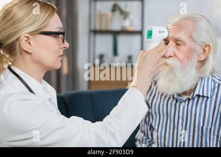 Nahaufnahme einer selbstbewussten medizinischen Mitarbeiterin, Messung der Körpertemperatur eines männlichen Patienten im Ruhestand zu Hause, mit einem berührungslosen Infrarot-Thermometer auf der Stirn. Coronavirus-Symptome. Stockfoto