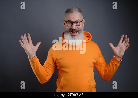 Reifer bärtiger Mann in Brillen gekleidet orange Hoodie lächelnd und Blick auf die Kamera auf einem grauen Hintergrund, der mit zwei Händen zeigt Nach oben Stockfoto