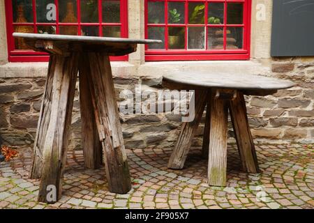 Alte runde Holztische auf Kopfsteinpflaster vor einem Steinmauer aus großen Steinen mit roten Holzfenstern Stockfoto