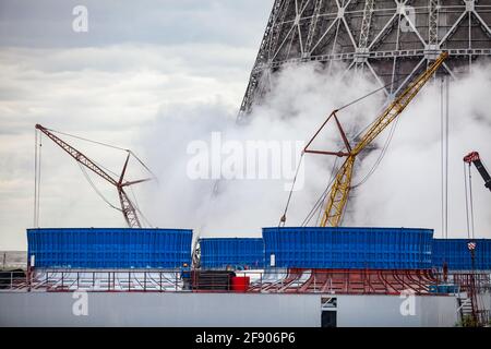 Elektroanlage beheizen. Bau neuer blauer kleiner Kühltürme. Großer Turm auf der Rückseite. Gelbe Mobilkrane, Dampf, graue Wolken. Karaganda, Kasachstan Stockfoto