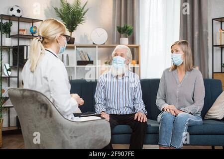 Rückansicht einer attraktiven Ärztin in Schutzmaske, die ihre Patienten besucht, eines angenehmen älteren Ehepaares in Masken zu Hause und dem Zuhören ihrer Symptome. Arzt und Patienten zu Hause während einer Pandemie Stockfoto