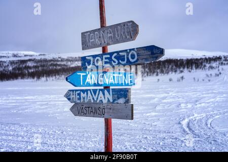 Hölzerne mehrfache Wegweiser zu den lokalen Dörfern in den Bergen Lapplands für Schneemobiltouristen. Sonniger Tag, verschneite Berge mit Birken an verschwommen Stockfoto