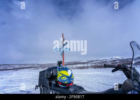 Hölzerner Wegweiser für Touristen zu Dörfern in den Bergen Lapplands. Verschwommenes Schneemobil und Helm im Vordergrund. Sonniger Tag, verschneite Berge Stockfoto