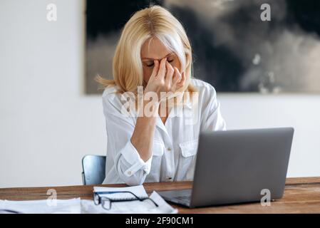 Müde verärgert kaukasische reife Blondine, ceo, Manager oder Freiberufler, in weißem Hemd, müde von Online-Arbeit auf Laptop, Stress erleben, Pause von der Arbeit, reibt ihre Augen mit der Hand, hat Kopfschmerzen Stockfoto