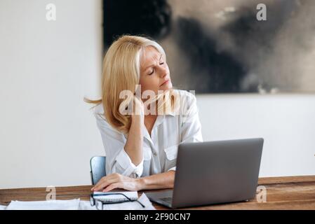 Erfolgreiche, entspannte, müde kaukasische blonde Frau mittleren Alters, Geschäftsfrau oder Anwalt, mit weißem Hemd, an einem Tisch sitzend, Pause nehmend, die Augen schließt, überarbeitet, erschöpft Stockfoto