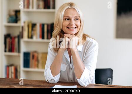 Porträt einer glücklichen, eleganten, erfolgreichen, reifen blonden Frau in weißem, stilvollem Hemd, sitzt am Schreibtisch im Büro, macht eine Pause von der Arbeit, blickt zur Seite, denkt, lächelt freundlich Stockfoto