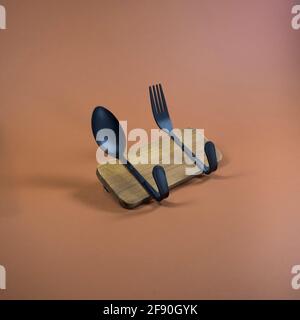 Küchenhaken aus Löffel und Gabel an Holzfliesen befestigt. Brauner, oranger Hintergrund. Minimale abstrakte Komposition. Essen, Getränke, Inneneinrichtung. Stockfoto