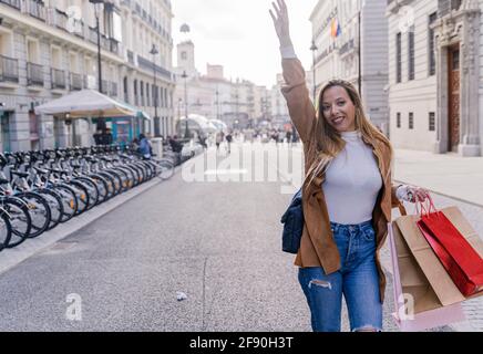 Lächelnde junge Touristenin wartet draußen auf ein Taxi. Fröhliche moderne Dame mit Tasche gestikuliert, um Signal an Taxifahrer zu geben. Taxi Service Konzept Stockfoto