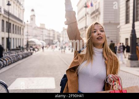 Lächelnde junge Touristenin wartet draußen auf ein Taxi. Fröhliche moderne Dame mit Tasche gestikuliert, um Signal an Taxifahrer zu geben. Taxi Service Konzept Stockfoto