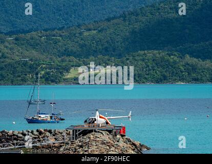 Airlie Beach, Queensland, Australien - 2021. April: Passagiere besteigen einen Hubschrauber auf einem Hubschrauberlandeplatz mit Booten im Hintergrund Stockfoto