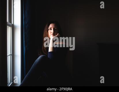 Attraktive Frau, die allein in einem dunklen Raum neben einem Fenster sitzt. Stockfoto