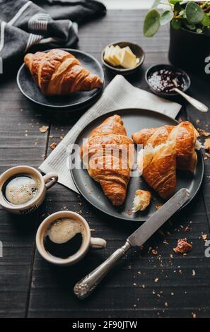 Teller mit Croissants mit Kaffee, Marmelade und Butter auf schwarzem Tisch. Stockfoto