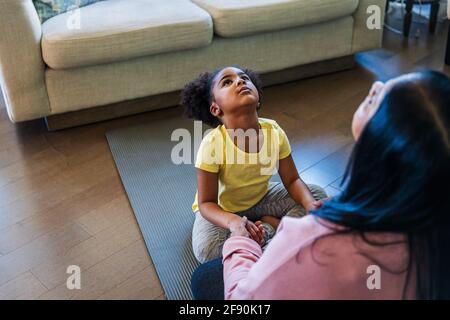 Mädchen, das mit Großmutter Yoga praktiziert, während sie auf einer Übungsmatte sitzt Im Wohnzimmer Stockfoto