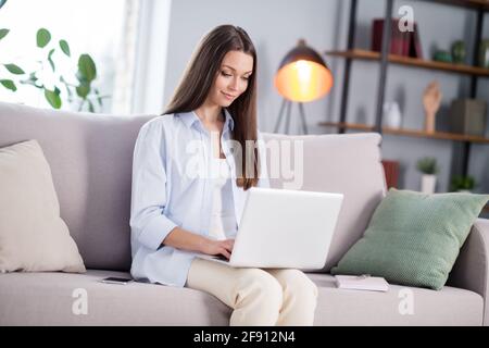 Foto in voller Größe von schönen optimistischen Brünette lange Frisur Dame Sitzen Sie mit Laptop Tragen Sie Hemd zu Hause Stockfoto