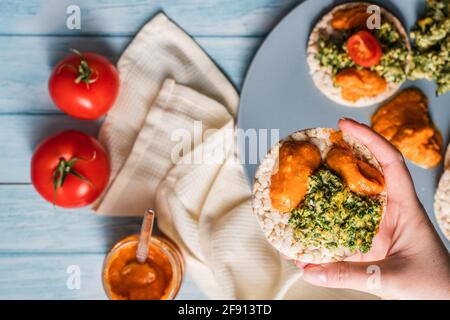 Knusprig aufgeblasene Reiskuchen auf dem Tisch mit Hummus Spread Spinat Mit Eiern und Tomaten und Gemüse auf dem Tisch - Draufsicht Hände halten an gesunden vege Stockfoto