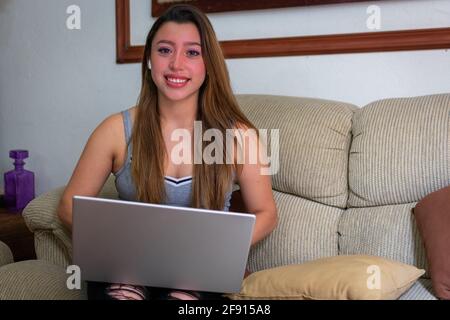 Die schöne junge Latina benutzt einen Laptop und lächelt im Sitzen Auf dem Sofa im Wohnzimmer Stockfoto