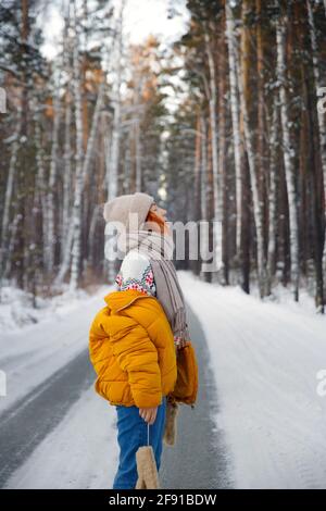 Das träumende rothaarige Mädchen steht mitten auf der Straße, ragt aus der Zunge, fängt Schneeflocken im Winterwald Stockfoto