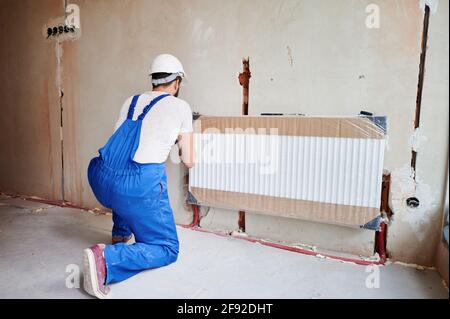 Zurück viow von Klempner, trägt blaue Overalls, weißer Helm, Installation von Wasserkühler in unvollendete Wohnung. Instandsetzungsarbeiten Renovierung in der Wohnung. Heizungsanlage. Horizontaler Schnappschuss Stockfoto