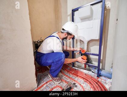 Klempner in Arbeit Overalls Installation verdeckter Toilettenrahmen im Badezimmer. Männlicher Arbeiter im Schutzhelm, der Clip auf die Rohrschelle setzt und ein an der Wand aufgehängtes Toilettensystem installiert. Konzept der Sanitär-Arbeiten. Stockfoto