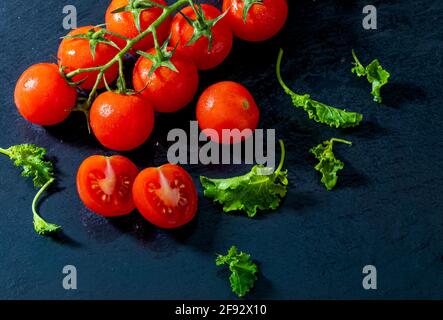 Grüne Blätter von Grünkohl und frische Weintomaten (Kirschen) mit Tropfen Wasser, roh, ganz und halbiert. Auf schwarzem texturierten Schieferhintergrund. Ingredie Stockfoto