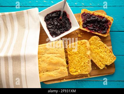 Maisbrot ist Brot mit Maismehl, Maismehl. Schöner, gelber Maisbrot-Laib mit Heidelbeermarmelade in einer Schüssel. Blauer Holzhintergrund. Flach liegend Stockfoto