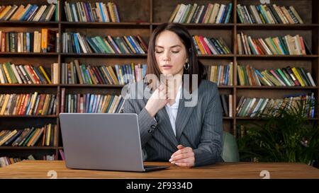 Professionelle Sekretärin in grauen Kostümtypen auf Laptop unter sitzen Starker Lüfterwind gegen verschiedenfarbige Bücher auf Racks in Büro Stockfoto