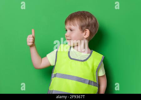 Kaukasischer Junge in hellen Farben helle Weste mit reflektierenden Streifen zeigt Daumen nach oben Geste, groß, und lächeln. Studio auf grünem Hintergrund, saf Stockfoto