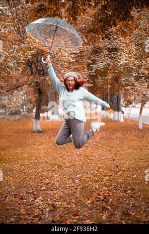Fröhliches kaukasisches Mädchen, das im Herbstpark mit einem transparenten Regenschirm aufspringt. Stockfoto