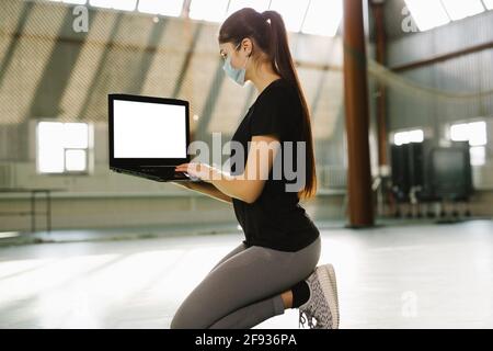 Der Fitnesstrainer kniet sich in einem leeren Fitnessstudio nieder. Frau in medizinischer Maske arbeitet entfernt in der Einsamkeit drinnen. Sie sitzt mit einem Laptop auf den Knien. Quarantäne sperren Stockfoto