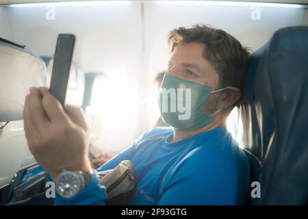 Reisen und Tourismus in Zeiten von Covid19 - Lifestyle-Portrait Von jungen attraktiven kaukasischen Mann in Gesichtsmaske mit mobilen Telefon im Flugzeug während des Fluges Stockfoto