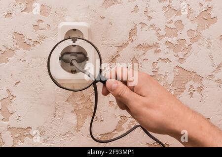 Die Hand eines Arbeiters mit einer Lupe untersucht ein Kabel, das an eine Doppelsteckdose angeschlossen ist. Das Konzept der Sicherheit von Haushaltsgeräten. Stockfoto