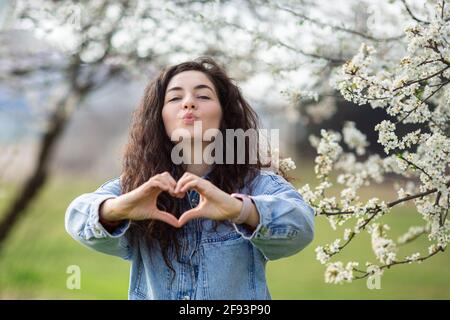 Schöne junge Frau, die mit ihren Händen ein Herz macht. Porträt eines lächelnden jungen Mädchens, das im Park posiert. Stockfoto