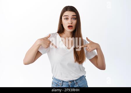 Überraschte Frau, die auf sich zeigt, die Augenbrauen hebt und erstaunt keuchelt, das Selbstförderungslogo zeigt, Banner vorführt, über Weiß stehend Stockfoto