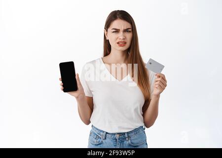 Enttäuscht runzelte weibliche Klientin, zeigte Smartphone und Kreditkarte, sieht frustriert aus, äußert Abneigung, beschwert sich über Online-Shopping-App Stockfoto