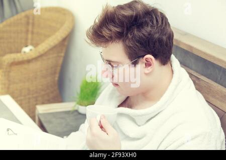Der junge Kaukasusmann liest in einem Bademantel mit einer Tasse Kaffee ein Buch. Am frühen Morgen im Bett. Stockfoto