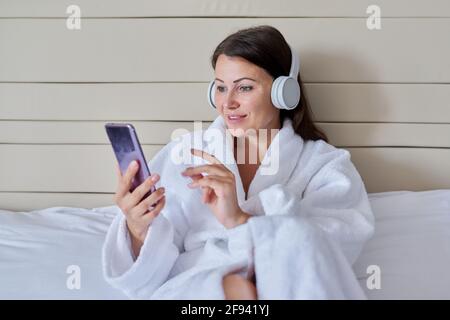 Glückliche reife Frau in weißem Bademantel und Kopfhörern mit Smartphone Sitzen im Bett Stockfoto